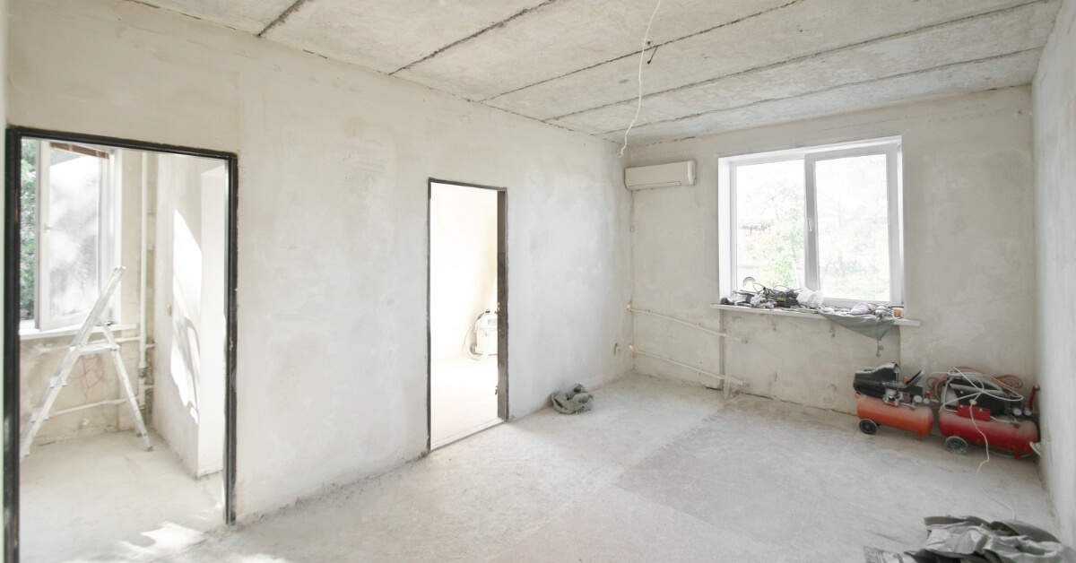 An empty room during an interior demolition phase in Ottawa, featuring bare concrete walls and floor. Construction tools, such as an air compressor and hand tools, are placed beside a window, reflecting the initial steps of a remodeling project.